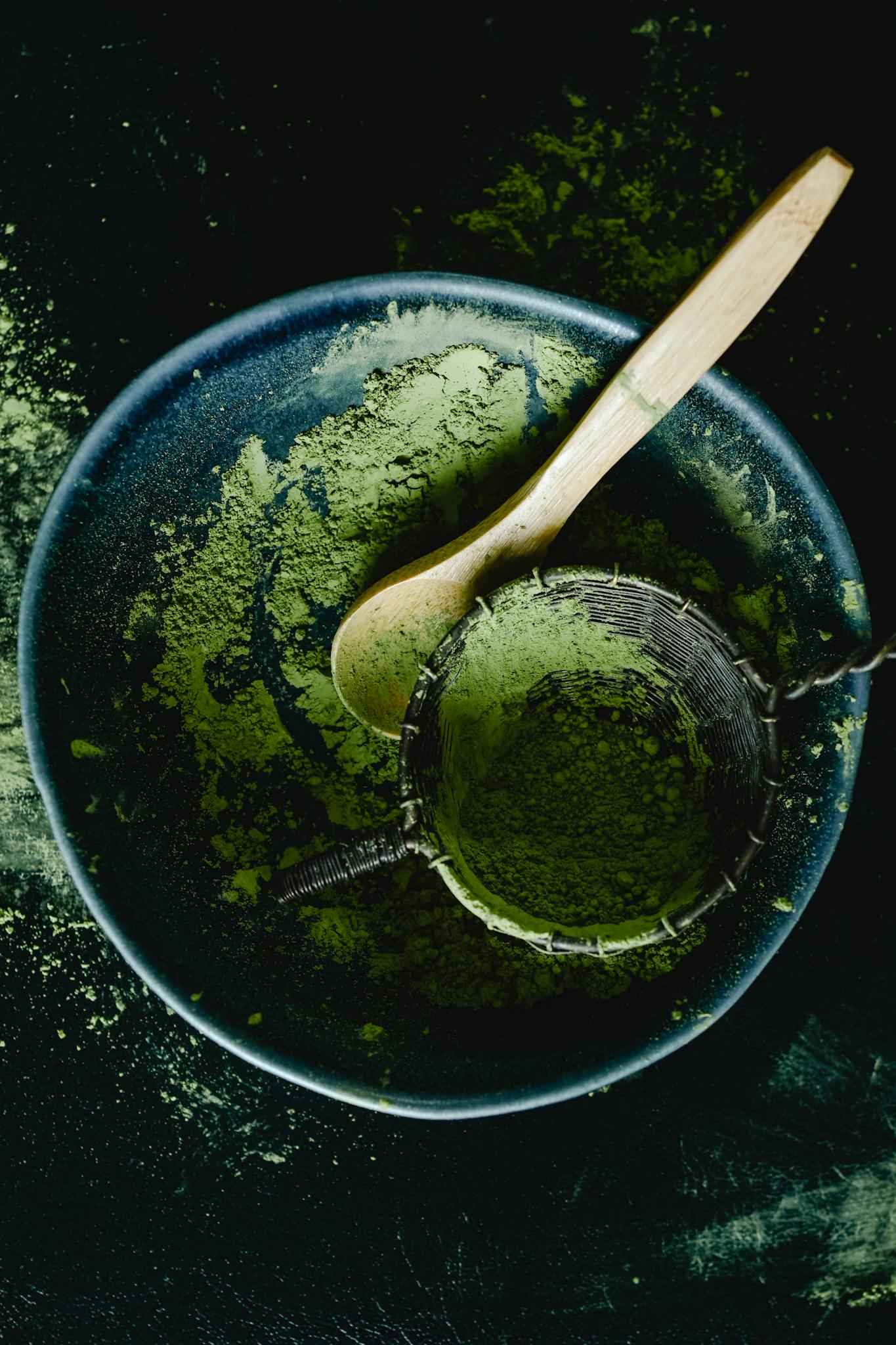 Overhead view of green matcha powder with a wooden spoon in a dark blue ceramic bowl, showcasing a rustic kitchen style.