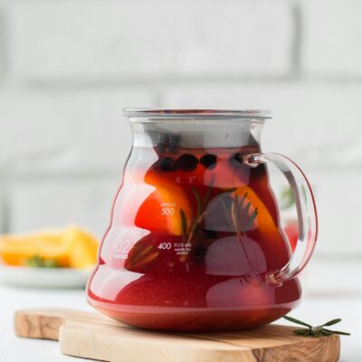 Refreshing homemade herbal tea with fruits and herbs in a glass teapot on a wooden board.