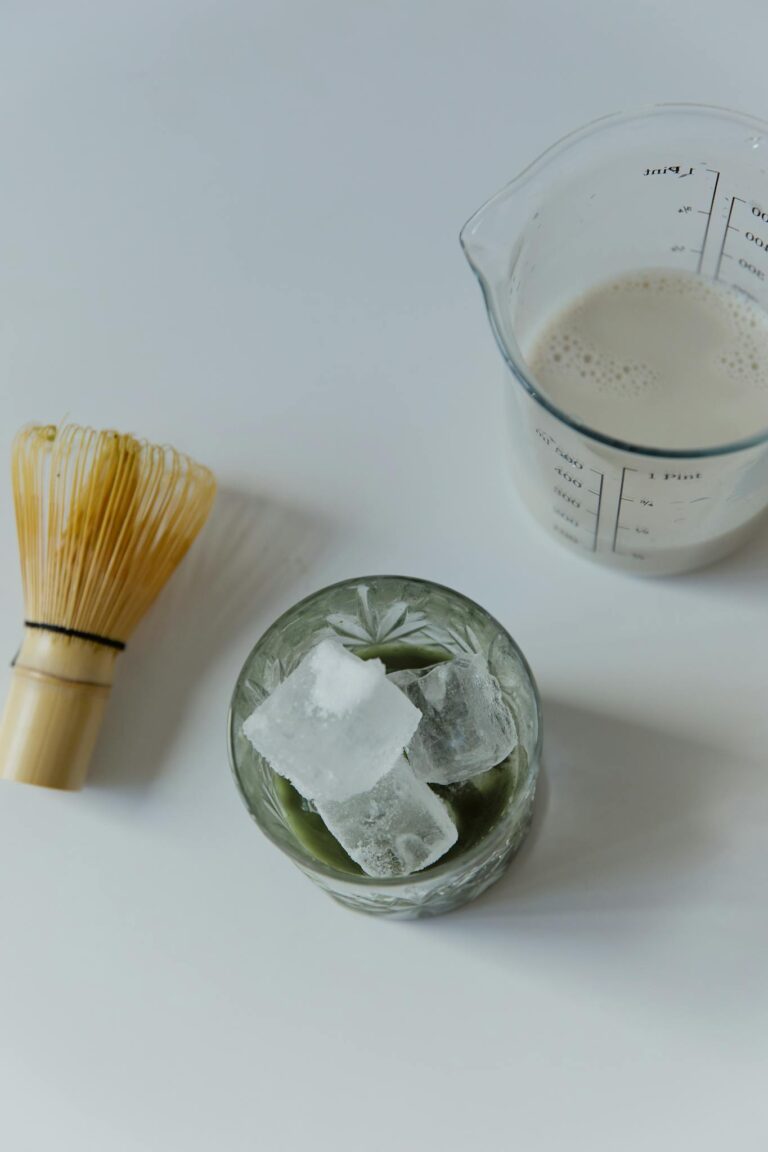 Top view of matcha latte ingredients including ice cubes, milk, and a whisk on a white background.